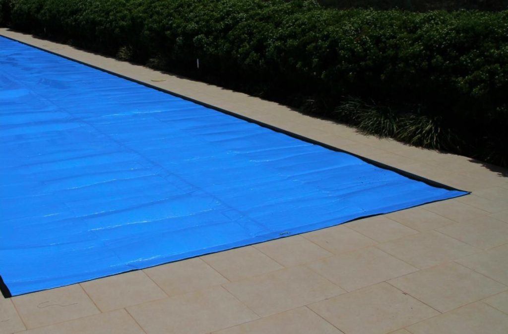 Blue solar pool cover laid on pavers beside a garden hedge