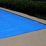 Blue solar pool cover laid on pavers beside a garden hedge