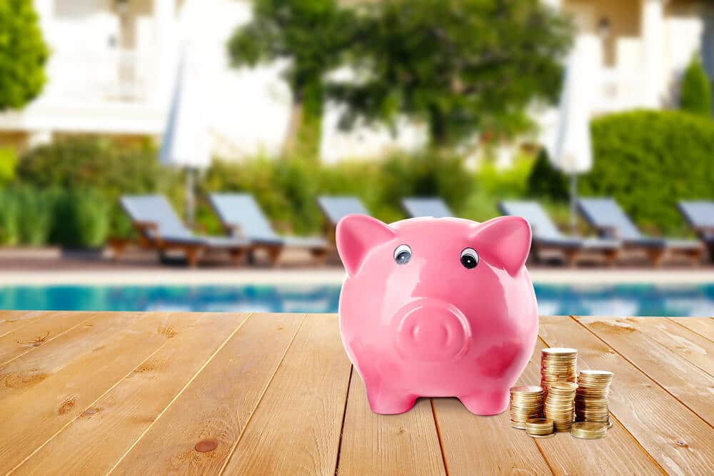 Pink piggy bank with stacked coins on a wooden poolside deck - The cheapest and fastest ways to heat a pool