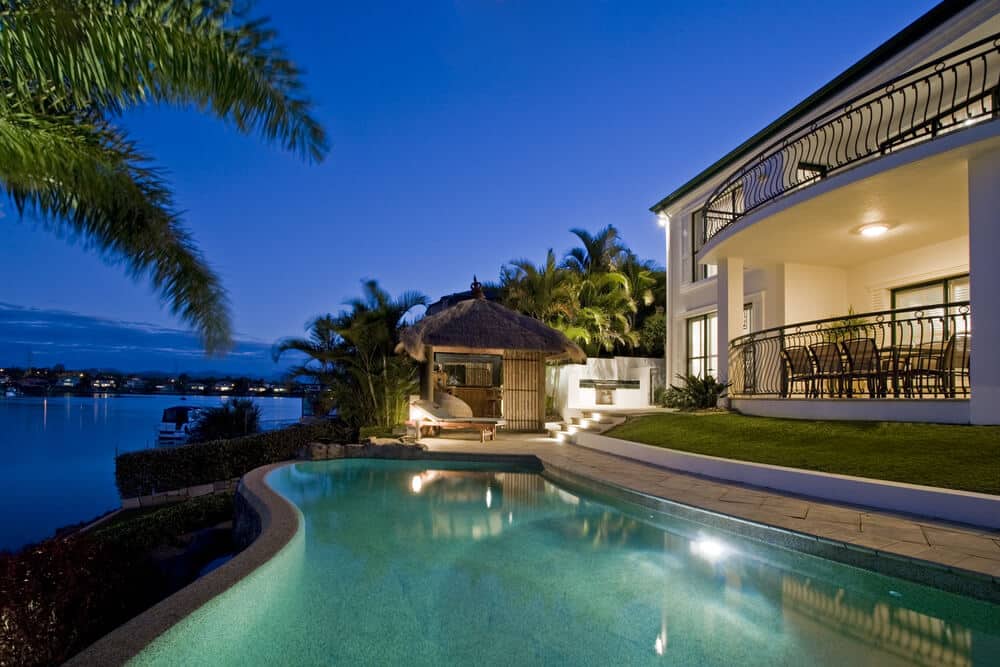 Luxury waterfront infinity pool lit up at night beside a mansion - Top 5 Tips for Installing a Pool Heater on the Gold Coast