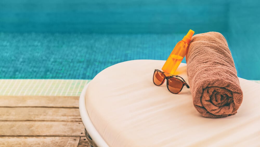 Sunglasses, sunscreen bottle and rolled towel on a poolside lounger - The cheapest way to heat your pool and keep summer going, longer!