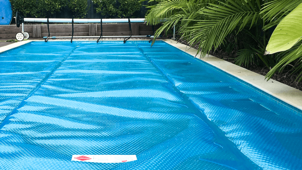 Blue thermal pool cover rolled across a backyard swimming pool surrounded by tropical plants